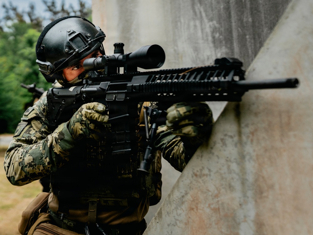 A tactical operator in full combat gear is positioned against a concrete wall in a shooting stance, aiming a rifle with an optical scope. The individual is equipped with military-grade protective equipment including a ballistic helmet, plate carrier vest, and camouflage uniform. The setting appears to be an outdoor training or operational environment.