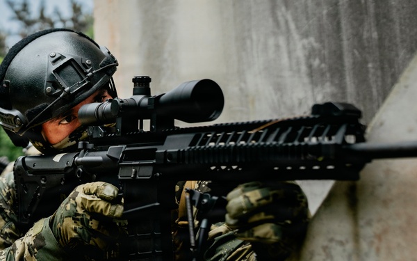 A tactical operator in full combat gear is positioned against a concrete wall in a shooting stance, aiming a rifle with an optical scope. The individual is equipped with military-grade protective equipment including a ballistic helmet, plate carrier vest, and camouflage uniform. The setting appears to be an outdoor training or operational environment.