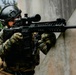 A tactical operator in full combat gear is positioned against a concrete wall in a shooting stance, aiming a rifle with an optical scope. The individual is equipped with military-grade protective equipment including a ballistic helmet, plate carrier vest, and camouflage uniform. The setting appears to be an outdoor training or operational environment.