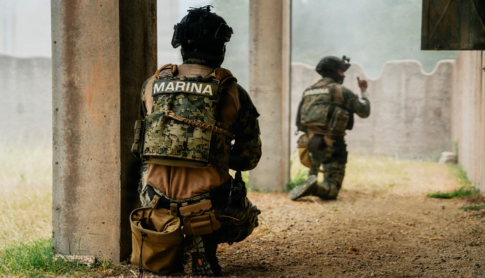 Two armed personnel wearing tactical gear marked 'MARINA' (Mexican Navy) are positioned in a kneeling stance near a concrete structure. The foreground operator is taking cover behind a pillar while the second operator ahead appears to be signaling with a hand gesture. The scene appears to depict a tactical training exercise or operational clearance procedure.