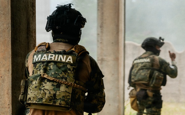 Two armed personnel wearing tactical gear marked 'MARINA' (Mexican Navy) are positioned in a kneeling stance near a concrete structure. The foreground operator is taking cover behind a pillar while the second operator ahead appears to be signaling with a hand gesture. The scene appears to depict a tactical training exercise or operational clearance procedure.