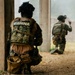Two armed personnel wearing tactical gear marked 'MARINA' (Mexican Navy) are positioned in a kneeling stance near a concrete structure. The foreground operator is taking cover behind a pillar while the second operator ahead appears to be signaling with a hand gesture. The scene appears to depict a tactical training exercise or operational clearance procedure.