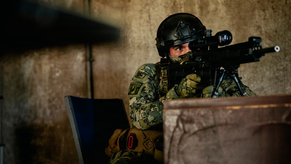 A uniformed military or special operations soldier wearing Mexican armed forces insignia is positioned behind cover, aiming a scoped rifle with a bipod. The soldier is equipped with full tactical gear including a ballistic helmet, face mask, camouflage uniform, and tactical gloves. The setting appears to be an indoor or semi-enclosed concrete structure.