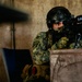A uniformed military or special operations soldier wearing Mexican armed forces insignia is positioned behind cover, aiming a scoped rifle with a bipod. The soldier is equipped with full tactical gear including a ballistic helmet, face mask, camouflage uniform, and tactical gloves. The setting appears to be an indoor or semi-enclosed concrete structure.