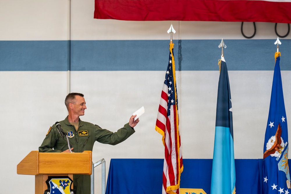 A military officer in olive green flight suit stands at a wooden podium during what appears to be a formal ceremony, holding a document and smiling. Three flags are visible behind him — the United States flag, a light blue flag (likely US Air Force), and a dark blue flag with a star (possibly a general officer's flag). The setting appears to be inside a military hangar or facility decorated for an official event.