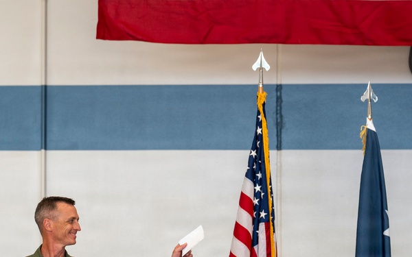 A military officer in olive green flight suit stands at a wooden podium during what appears to be a formal ceremony, holding a document and smiling. Three flags are visible behind him — the United States flag, a light blue flag (likely US Air Force), and a dark blue flag with a star (possibly a general officer's flag). The setting appears to be inside a military hangar or facility decorated for an official event.