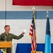 A military officer in olive green flight suit stands at a wooden podium during what appears to be a formal ceremony, holding a document and smiling. Three flags are visible behind him — the United States flag, a light blue flag (likely US Air Force), and a dark blue flag with a star (possibly a general officer's flag). The setting appears to be inside a military hangar or facility decorated for an official event.
