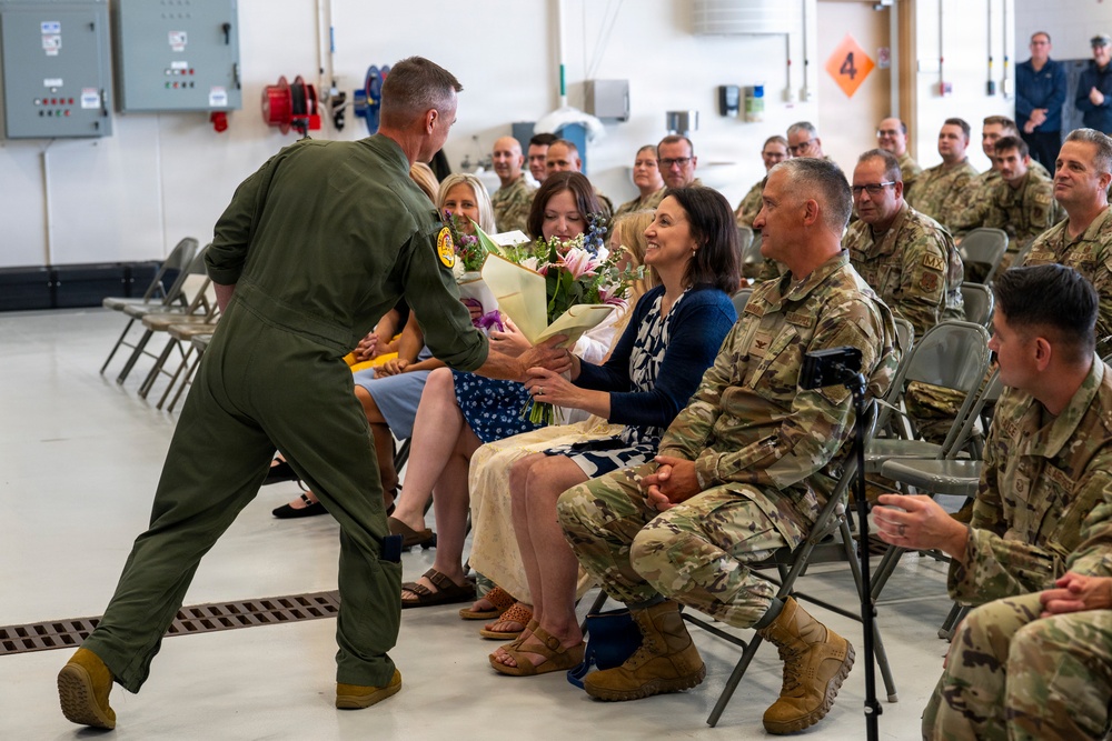 A military ceremony taking place inside what appears to be a hangar or large indoor facility. A person in an olive green flight suit is presenting a bouquet of flowers to a seated civilian woman, surrounded by rows of military personnel in camouflage uniforms. The atmosphere appears celebratory, consistent with a change of command, retirement, or recognition ceremony.
