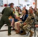 A military ceremony taking place inside what appears to be a hangar or large indoor facility. A person in an olive green flight suit is presenting a bouquet of flowers to a seated civilian woman, surrounded by rows of military personnel in camouflage uniforms. The atmosphere appears celebratory, consistent with a change of command, retirement, or recognition ceremony.