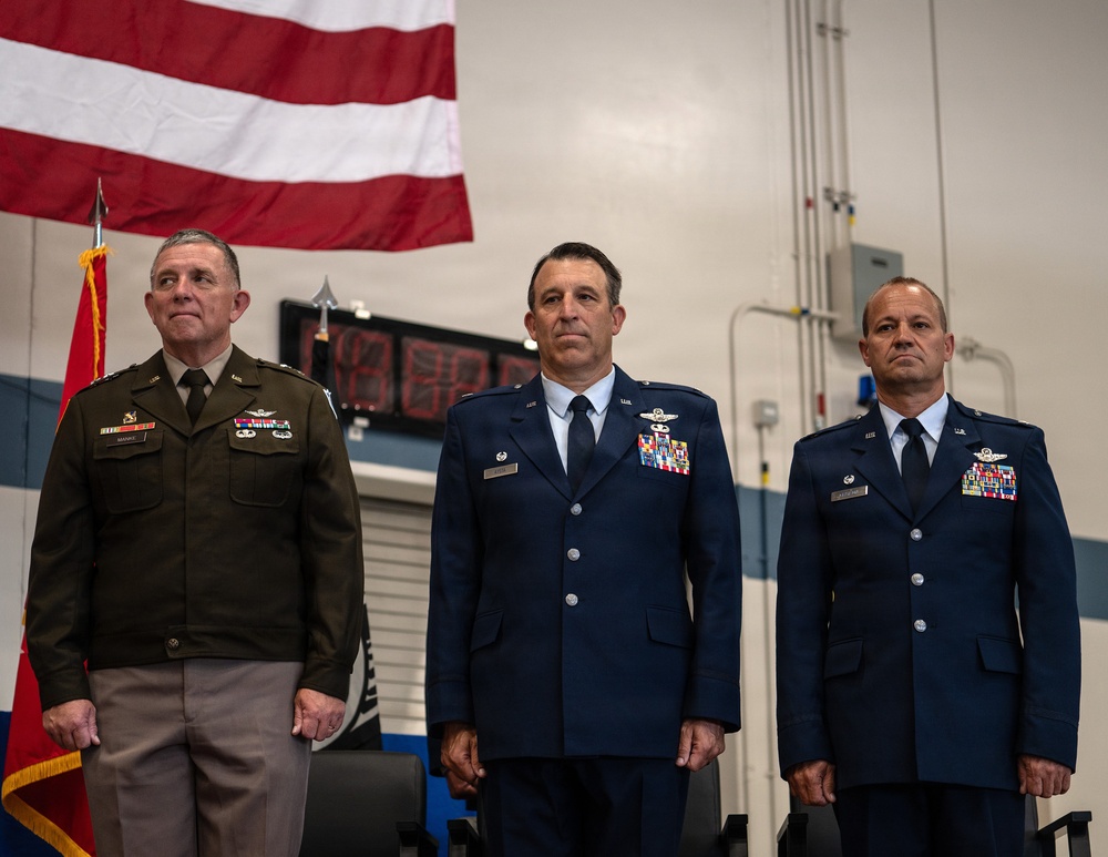 Three senior military officers stand at attention during a formal military ceremony, likely a change of command or awards ceremony. The officer on the left wears a U.S. Army dress uniform in olive green, while the two officers on the right wear U.S. Air Force dress blue uniforms with extensive medal and ribbon displays. A large American flag is prominently displayed in the background.