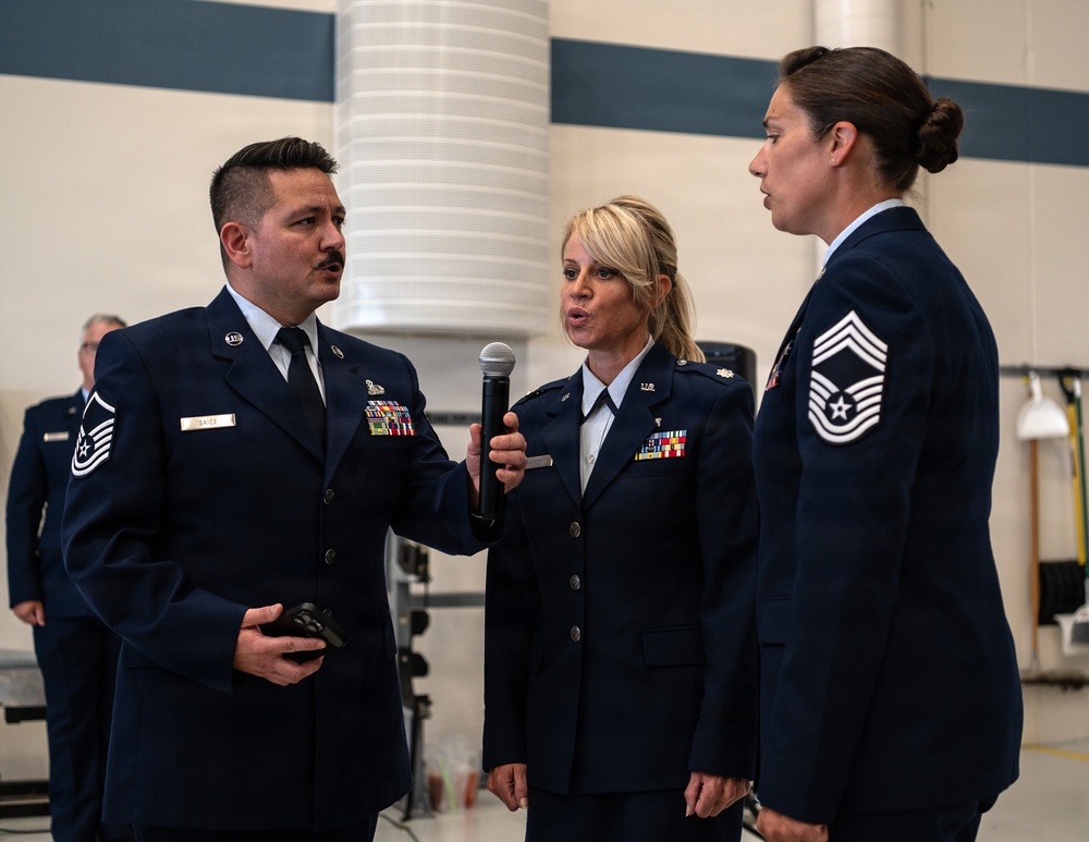 Three U.S. Air Force personnel in dress blue uniforms are engaged in what appears to be a formal military ceremony or event inside a hangar facility. A female officer in the center is holding a microphone and speaking, while a male Senior Master Sergeant on the left and a female Chief Master Sergeant on the right are listening. The setting suggests an official recognition, promotion, or change of command ceremony.