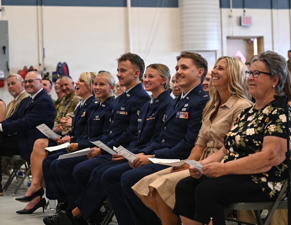 A group of uniformed military personnel and civilians are seated in rows at what appears to be an official ceremony held inside a hangar or large indoor facility. Three individuals in dark blue military dress uniforms with ribbons and insignia are prominently featured in the foreground, smiling and holding printed programs. They are surrounded by family members or guests in civilian attire who are also smiling and laughing.