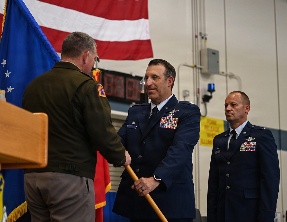A military ceremony is taking place indoors, likely a change of command or award presentation. A U.S. Air Force officer in dress blue uniform is receiving or exchanging a guidon (ceremonial staff) from an Army officer in green service uniform. A second Air Force officer stands at attention in the background.