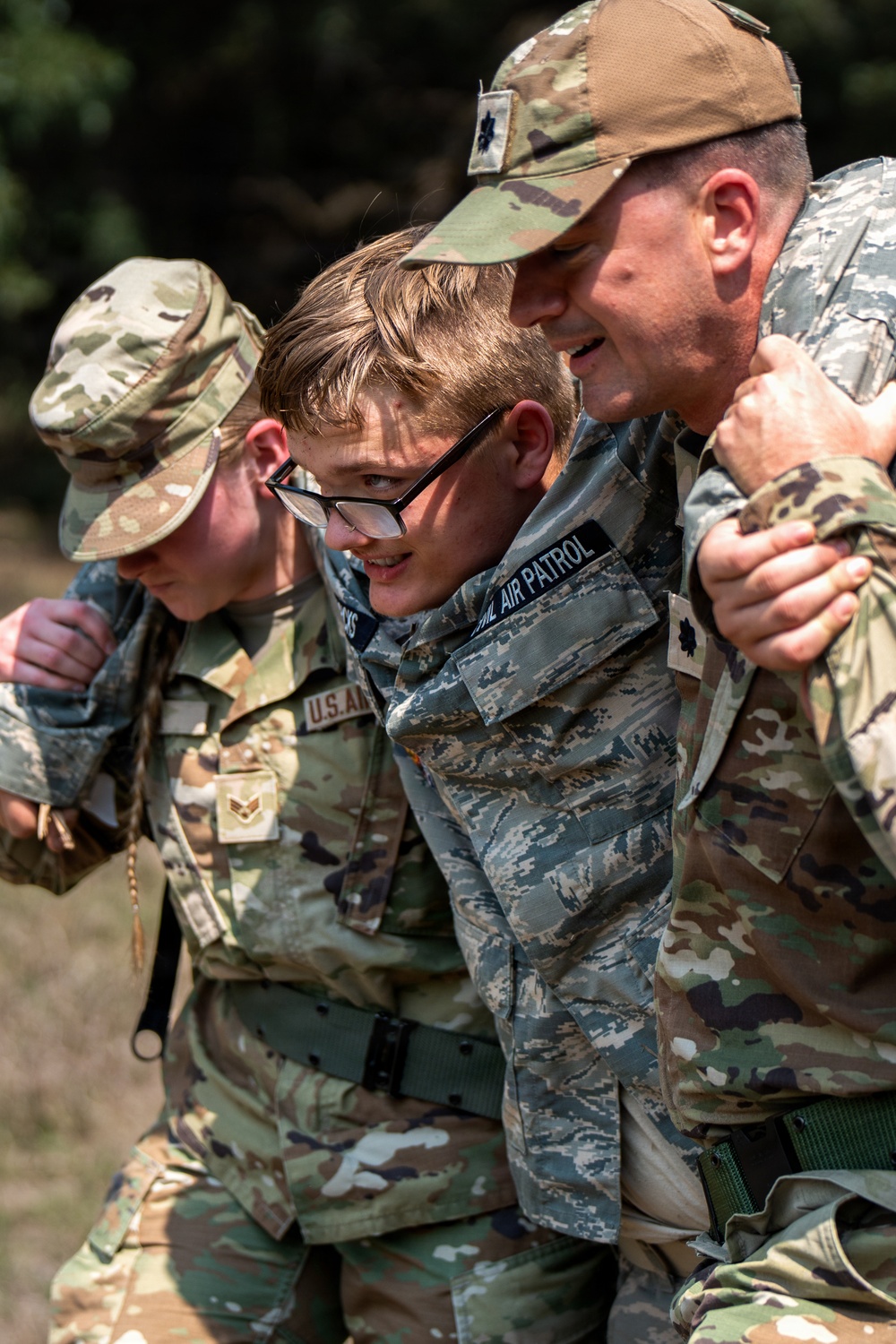 Three individuals in military uniforms are engaged in what appears to be a casualty assistance or buddy-carry exercise. The central figure, wearing a Civil Air Patrol uniform, is being supported on both sides by two others in OCP (Operational Camouflage Pattern) uniforms. The scene suggests a training exercise focused on personnel recovery or first aid procedures.