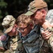 Three individuals in military uniforms are engaged in what appears to be a casualty assistance or buddy-carry exercise. The central figure, wearing a Civil Air Patrol uniform, is being supported on both sides by two others in OCP (Operational Camouflage Pattern) uniforms. The scene suggests a training exercise focused on personnel recovery or first aid procedures.