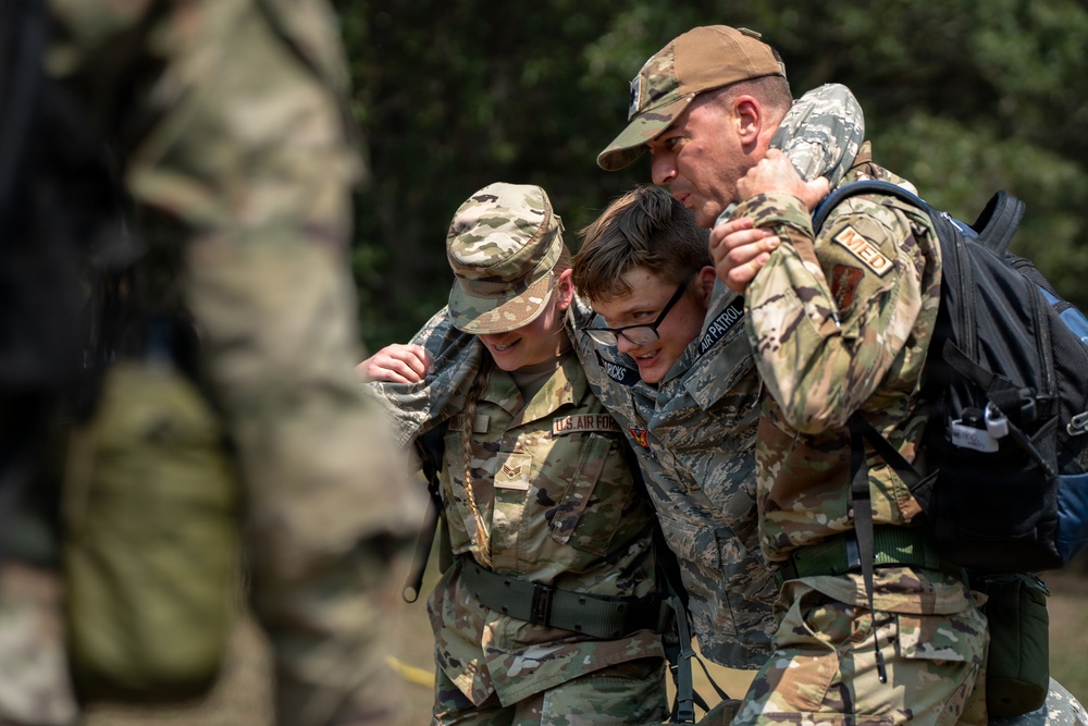 Three individuals in military uniforms are assisting or supporting a younger person wearing glasses who appears to be a Civil Air Patrol cadet. Two adults in camouflage uniforms — one wearing a brown cap with a 'MED' patch and carrying a backpack — are helping support the cadet between them. The scene appears to take place outdoors during a training exercise or field activity.