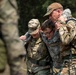 Three individuals in military uniforms are assisting or supporting a younger person wearing glasses who appears to be a Civil Air Patrol cadet. Two adults in camouflage uniforms — one wearing a brown cap with a 'MED' patch and carrying a backpack — are helping support the cadet between them. The scene appears to take place outdoors during a training exercise or field activity.