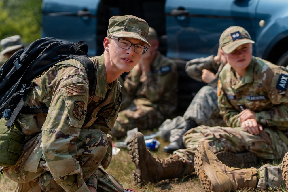 The image shows several young individuals wearing OCP (Operational Camouflage Pattern) uniforms with Civil Air Patrol insignia, resting on the ground near a vehicle. The foreground subject is wearing glasses and carrying a black backpack, looking directly at the camera. The group appears to be taking a break during a training exercise or field activity.