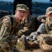 The image shows several young individuals wearing OCP (Operational Camouflage Pattern) uniforms with Civil Air Patrol insignia, resting on the ground near a vehicle. The foreground subject is wearing glasses and carrying a black backpack, looking directly at the camera. The group appears to be taking a break during a training exercise or field activity.