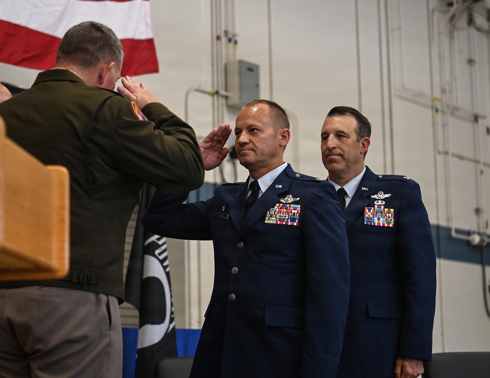 A military ceremony is taking place inside what appears to be a hangar or large indoor facility. Two U.S. Air Force officers in dress blue uniforms are standing at attention while a third individual in an olive/green flight suit is rendering a salute. An American flag and a POW/MIA flag are visible in the background, suggesting this may be a change of command or awards ceremony.