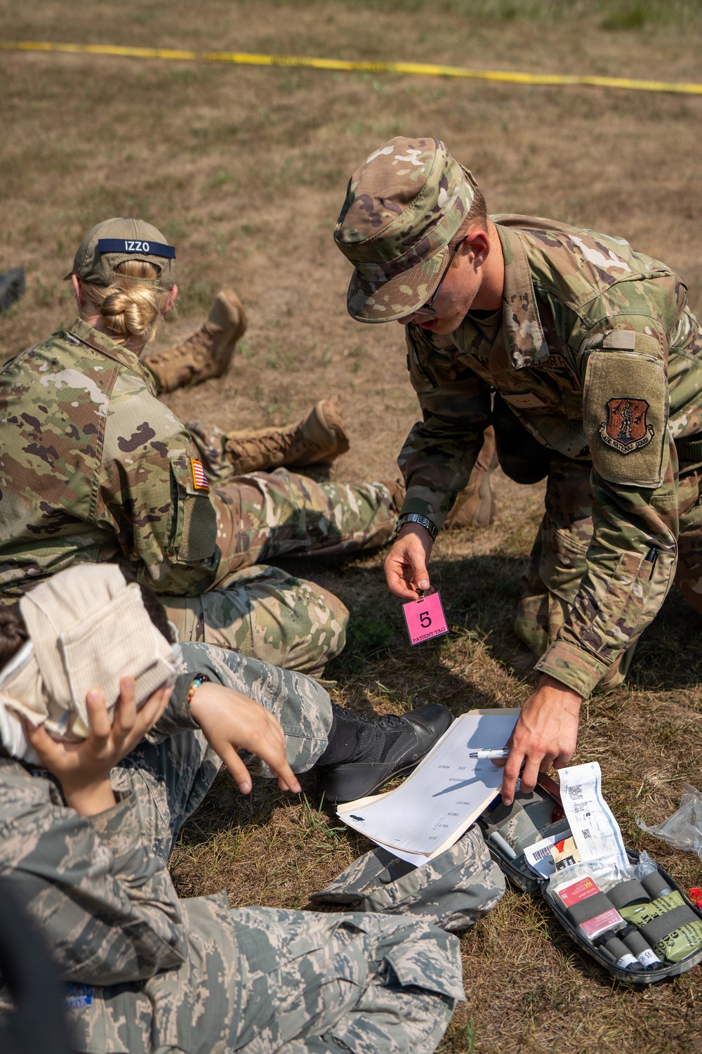 Military personnel in OCP (Operational Camouflage Pattern) uniforms are conducting a field medical training exercise. A service member is holding a pink 'Patient Tag' numbered '5' while referencing paperwork and a medical kit on the ground. At least one simulated casualty is lying on the ground being assessed by the team.