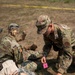 Military personnel in OCP (Operational Camouflage Pattern) uniforms are conducting a field medical training exercise. A service member is holding a pink 'Patient Tag' numbered '5' while referencing paperwork and a medical kit on the ground. At least one simulated casualty is lying on the ground being assessed by the team.