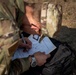 A military personnel member in MultiCam/OCP camouflage uniform is writing on a form that contains a human body diagram, likely a medical injury assessment or casualty card. The individual is seated outdoors on dry ground, using a black backpack as a writing surface. A name tape reading 'MASON' is visible on the uniform.