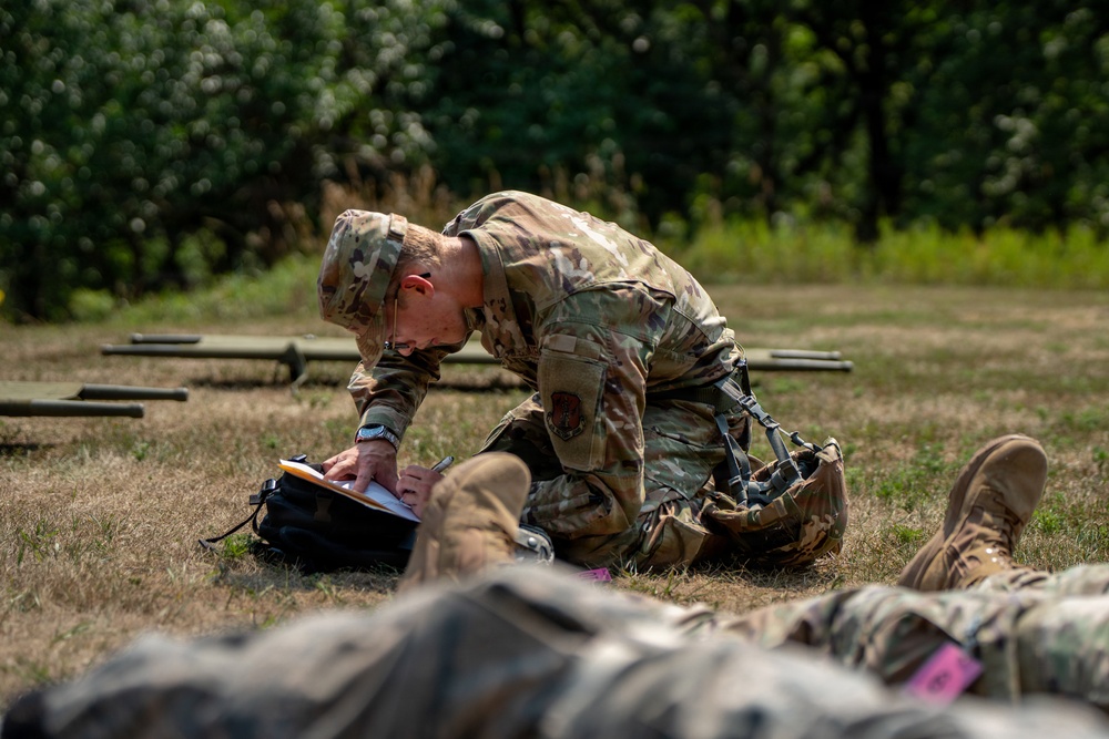 A soldier in OCP (Operational Camouflage Pattern) uniform is kneeling on grass and writing on a clipboard or notepad. In the foreground, another person in camouflage uniform is lying on the ground, possibly simulating a casualty in a training exercise. The setting appears to be an outdoor military training area with wooden obstacles visible in the background.