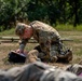 A soldier in OCP (Operational Camouflage Pattern) uniform is kneeling on grass and writing on a clipboard or notepad. In the foreground, another person in camouflage uniform is lying on the ground, possibly simulating a casualty in a training exercise. The setting appears to be an outdoor military training area with wooden obstacles visible in the background.