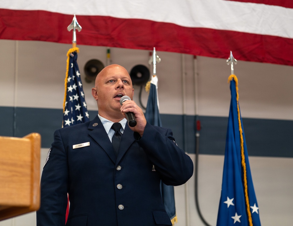 A U.S. Air Force enlisted member, identified by name tag as 'HINKS', is speaking into a handheld microphone at what appears to be an official military ceremony. He is wearing the Air Force Service Dress uniform and is positioned in front of multiple flags including the U.S. flag and Air Force flag. The setting appears to be an indoor military facility or hangar decorated for a formal event.