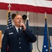 A U.S. Air Force enlisted member, identified by name tag as 'HINKS', is speaking into a handheld microphone at what appears to be an official military ceremony. He is wearing the Air Force Service Dress uniform and is positioned in front of multiple flags including the U.S. flag and Air Force flag. The setting appears to be an indoor military facility or hangar decorated for a formal event.