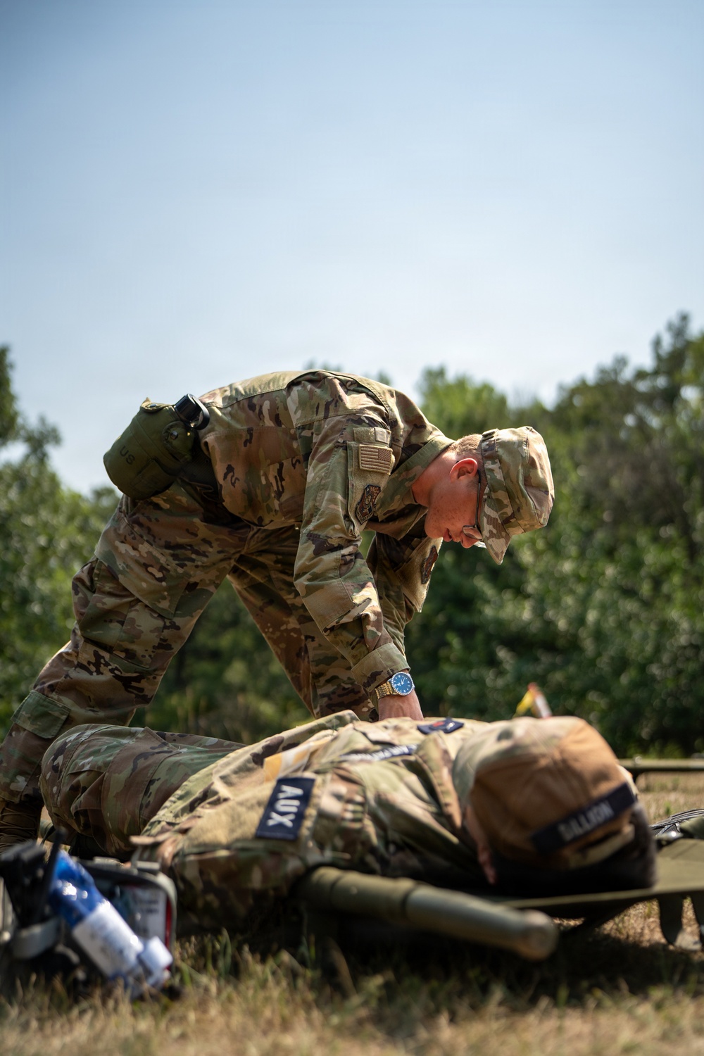 A soldier in OCP (Operational Camouflage Pattern) uniform is leaning over and attending to another soldier lying on a military litter/stretcher on the ground. The standing soldier appears to be performing a medical assessment or treatment on the prone individual during what appears to be a field training exercise. Medical supplies are visible nearby, and both individuals wear military uniforms with visible patches.