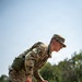 A U.S. Air Force service member in OCP (Operational Camouflage Pattern) uniform is leaning over another person lying on the ground, appearing to be conducting a casualty assessment or combat lifesaver drill. The standing individual wears glasses, a patrol cap, a wristwatch, and has a unit patch and U.S. flag patch visible on the left sleeve. The setting appears to be an outdoor training environment with trees in the background.