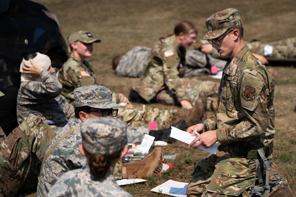 A group of military personnel in camouflage uniforms are engaged in what appears to be a field medical training exercise outdoors. One individual in OCP (Operational Camouflage Pattern) uniform is standing and reviewing paperwork while others practice bandaging and first aid techniques on the ground. The scene suggests a structured training scenario with multiple participants in various stages of casualty care practice.