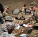 A group of military personnel in camouflage uniforms are engaged in what appears to be a field medical training exercise outdoors. One individual in OCP (Operational Camouflage Pattern) uniform is standing and reviewing paperwork while others practice bandaging and first aid techniques on the ground. The scene suggests a structured training scenario with multiple participants in various stages of casualty care practice.