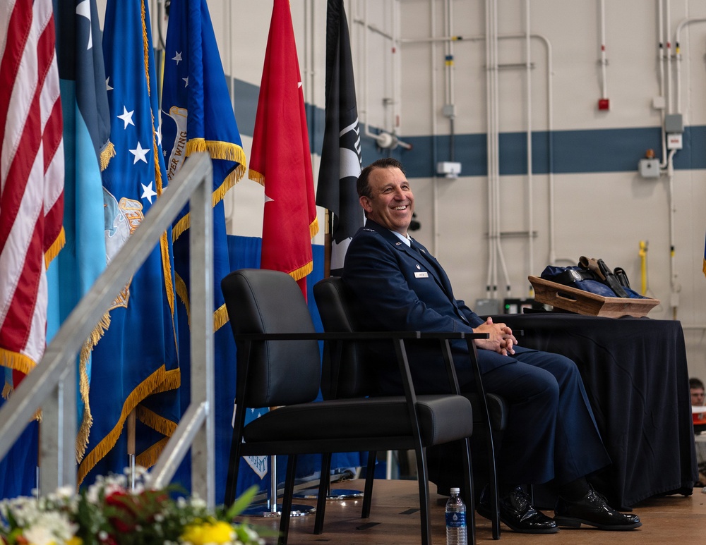 A military officer in a dark blue Air Force dress uniform is seated on a stage, smiling broadly during what appears to be a formal military ceremony. Multiple flags are displayed behind him, including the U.S. flag, Air Force flag, and POW/MIA flag. A table with items including what appear to be shoes or boots on a wooden tray is visible to his right.
