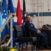 A military officer in a dark blue Air Force dress uniform is seated on a stage, smiling broadly during what appears to be a formal military ceremony. Multiple flags are displayed behind him, including the U.S. flag, Air Force flag, and POW/MIA flag. A table with items including what appear to be shoes or boots on a wooden tray is visible to his right.