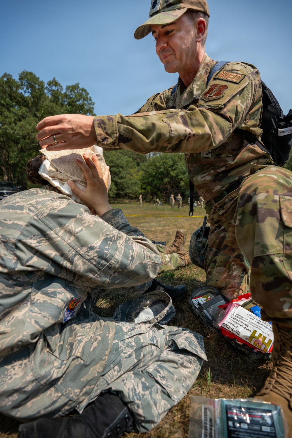 A military medic wearing OCP (Operational Camouflage Pattern) uniform with a 'MED' patch is providing first aid to another service member in ABU (Airman Battle Uniform) during what appears to be a field training exercise. The medic is applying a bandage or dressing to the seated casualty's head area. Medical supplies are visible on the ground nearby, suggesting a simulated casualty care scenario.