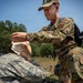 A military medic wearing OCP (Operational Camouflage Pattern) uniform with a 'MED' patch is providing first aid to another service member in ABU (Airman Battle Uniform) during what appears to be a field training exercise. The medic is applying a bandage or dressing to the seated casualty's head area. Medical supplies are visible on the ground nearby, suggesting a simulated casualty care scenario.