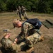 Military personnel are conducting a field medical training exercise in an outdoor grassy area. A medic wearing a 'MED' patch and carrying a backpack is kneeling and administering treatment to a seated soldier identified by a 'IZZO' name tape, while another person in Air Force ABU (Airman Battle Uniform) lies on the ground nearby with a medical kit open. Additional uniformed personnel are visible in the background approaching the scene.