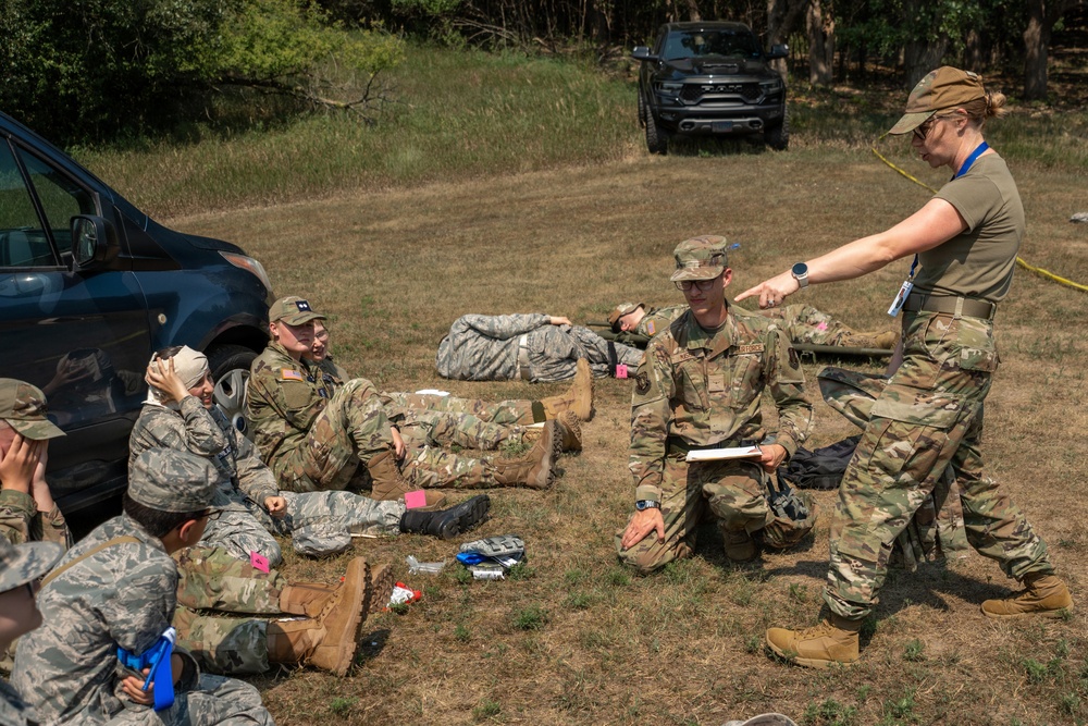 Military personnel in camouflage uniforms are engaged in what appears to be a field training exercise, likely a tactical combat casualty care (TCCC) or mass casualty triage drill. Several individuals are positioned on the ground simulating casualties, while an instructor or evaluator wearing a blue lanyard badge points and provides direction to a kneeling service member holding a clipboard. The setting is an outdoor grassy area with vehicles and trees in the background.