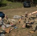 Military personnel in camouflage uniforms are engaged in what appears to be a field training exercise, likely a tactical combat casualty care (TCCC) or mass casualty triage drill. Several individuals are positioned on the ground simulating casualties, while an instructor or evaluator wearing a blue lanyard badge points and provides direction to a kneeling service member holding a clipboard. The setting is an outdoor grassy area with vehicles and trees in the background.