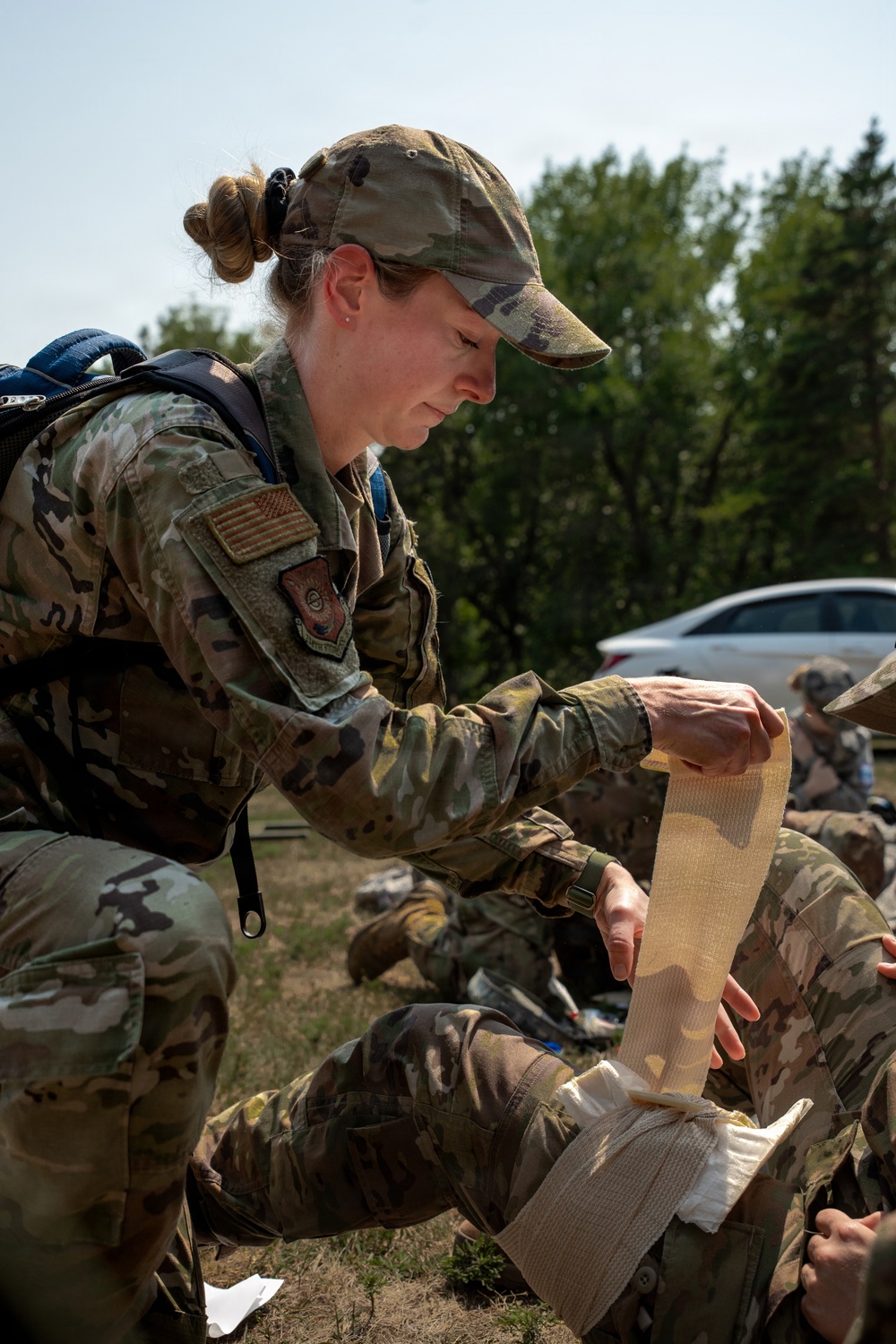 A female U.S. military service member in OCP (Operational Camouflage Pattern) uniform is applying a bandage or pressure dressing to another person's limb during what appears to be a field medical training exercise. She is wearing a camouflage cap, has a backpack, and displays a U.S. flag patch on her right shoulder. The outdoor setting with other personnel visible in the background suggests a group training scenario.