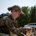 A female U.S. military service member in OCP (Operational Camouflage Pattern) uniform is applying a bandage or pressure dressing to another person's limb during what appears to be a field medical training exercise. She is wearing a camouflage cap, has a backpack, and displays a U.S. flag patch on her right shoulder. The outdoor setting with other personnel visible in the background suggests a group training scenario.