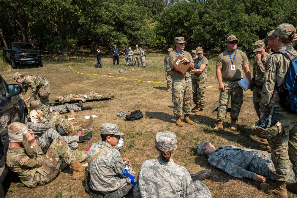 A military field training exercise depicting a mass casualty scenario with personnel in various uniforms acting as both casualties and responders. Evaluators wearing blue lanyards/badges observe and assess participants performing triage and medical response procedures. The exercise takes place in an outdoor grassy area bordered by trees, with yellow caution tape marking the scene perimeter.