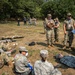 A military field training exercise depicting a mass casualty scenario with personnel in various uniforms acting as both casualties and responders. Evaluators wearing blue lanyards/badges observe and assess participants performing triage and medical response procedures. The exercise takes place in an outdoor grassy area bordered by trees, with yellow caution tape marking the scene perimeter.