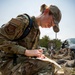 A female military service member in OCP (Operational Camouflage Pattern) uniform is kneeling on the ground and writing on a document or map. She is wearing a unit patch on her left shoulder and a name tape reading 'DOWELL,' and carries a hydration pack. In the background, a group of similarly uniformed personnel are seated on the ground in a field setting.