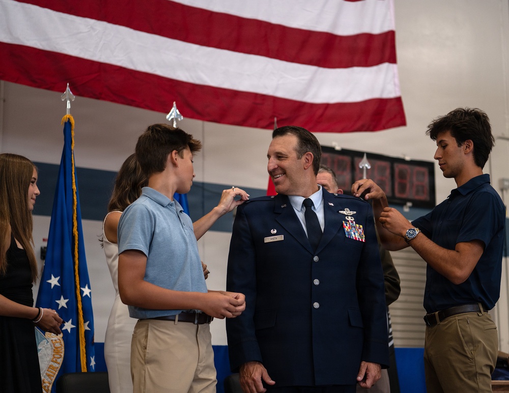 A U.S. Air Force officer is being pinned with new rank insignia on his shoulders by what appear to be family members, likely his children, during a formal promotion ceremony. The officer is smiling broadly while two young men pin his shoulders simultaneously, a traditional military promotion ritual. The ceremony takes place in front of a large American flag and an Air Force flag.