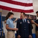 A U.S. Air Force officer is being pinned with new rank insignia on his shoulders by what appear to be family members, likely his children, during a formal promotion ceremony. The officer is smiling broadly while two young men pin his shoulders simultaneously, a traditional military promotion ritual. The ceremony takes place in front of a large American flag and an Air Force flag.