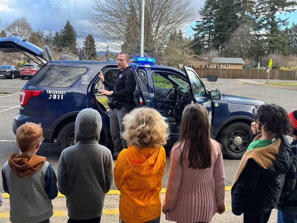 A police officer is conducting an educational demonstration for a group of children in a parking lot, showing them a police patrol SUV. The officer is standing beside the open door of a dark navy blue police vehicle with its emergency lights activated. This appears to be a community outreach or school visit event where children are learning about law enforcement.